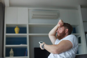 A bearded man of European descent holds his hand up to his head as he struggles indoors with no AC.